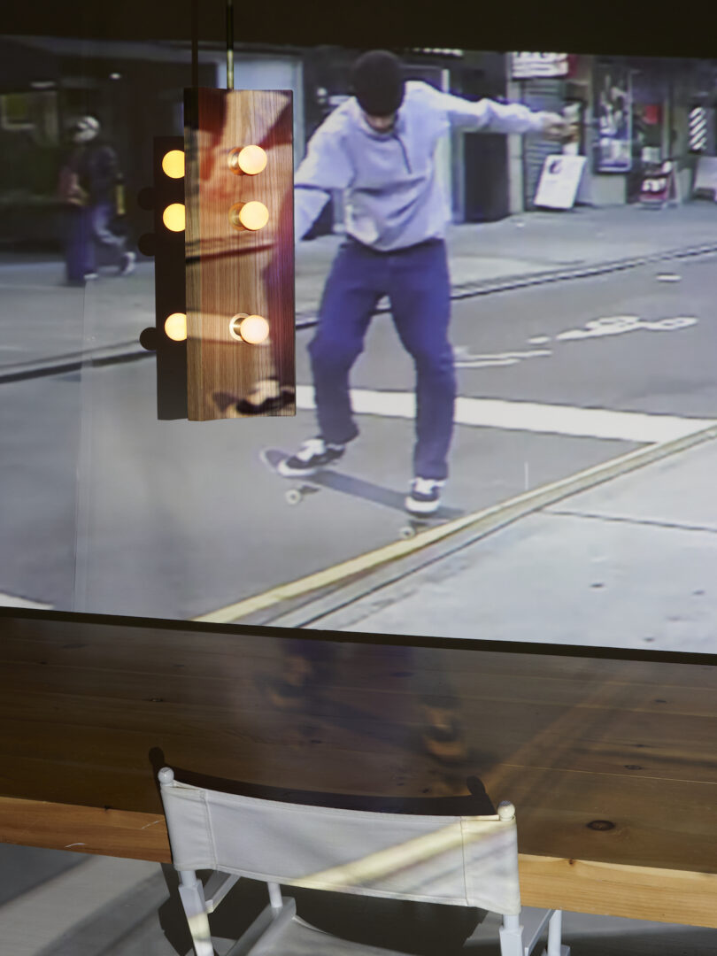 Projection of a skateboarder performing a trick on a city street, with a visible streetlamp. A table and chair are in the foreground, illuminated by overhead lights.