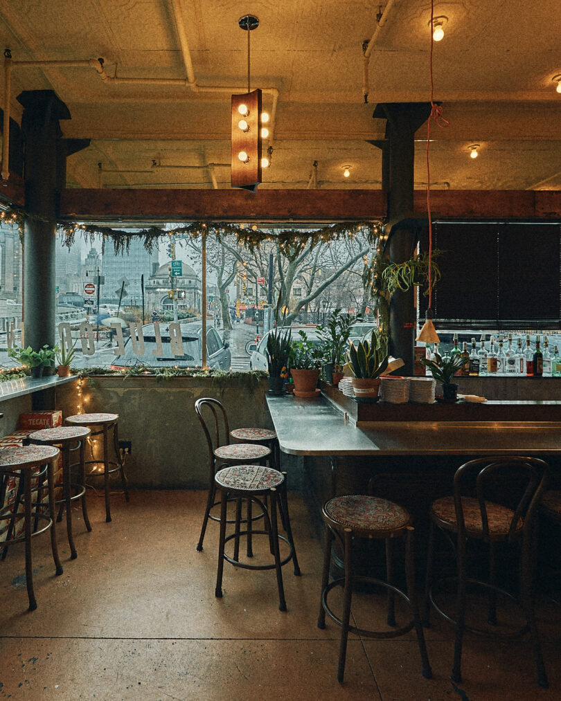 Cozy bar interior with wooden stools around tables, plants on the counter, and a large window overlooking a street scene. Dim lighting adds warmth to the setting