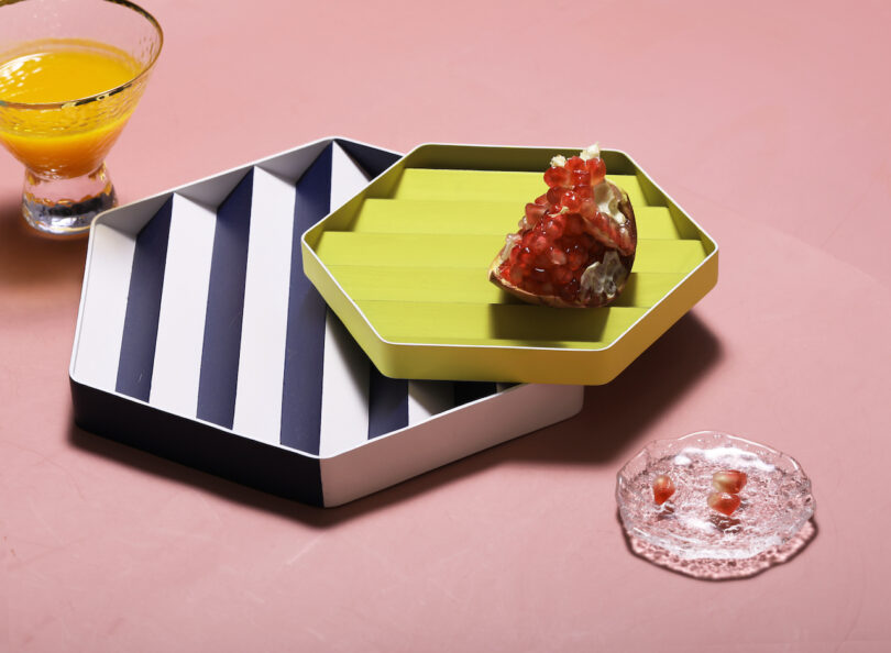 A hexagonal tray with green and black stripes holds a pomegranate half. A glass of orange juice and spilled ice are nearby on a pink surface