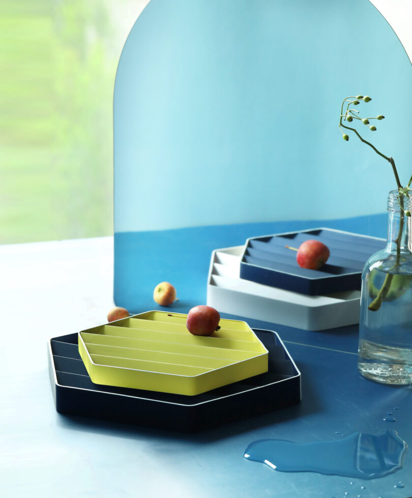 Stacked hexagonal trays with fruits sit on a blue surface, next to a vase with small branches. A spilled liquid is nearby