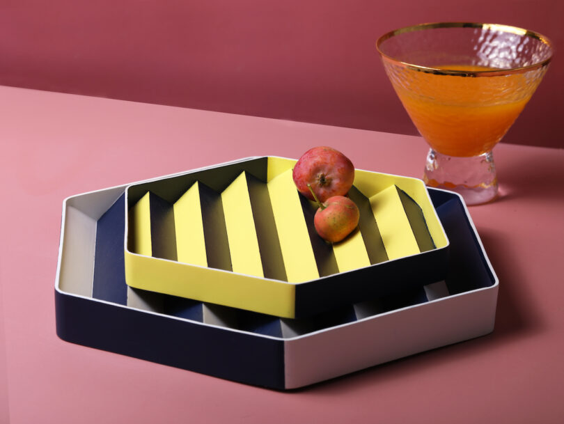 Two apples on yellow striped trays, with a glass of orange juice on a pink surface