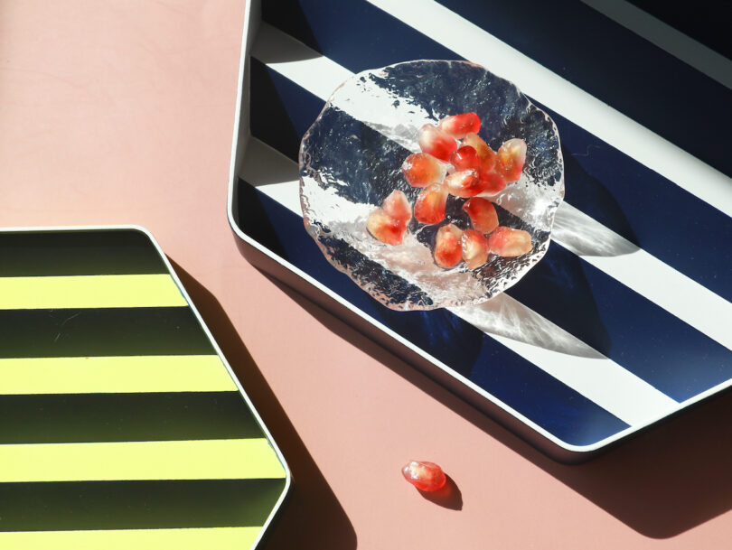 Glass plate with pomegranate seeds on a blue and white striped tray, next to a black and yellow tray, on a pink surface