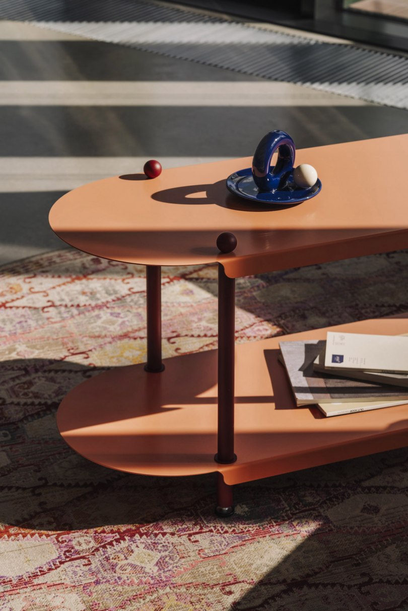 A modern coffee table with a two-tier orange design. The top tier holds a blue decorative item and a small ball. The lower tier has a stack of papers. A patterned rug is underneath