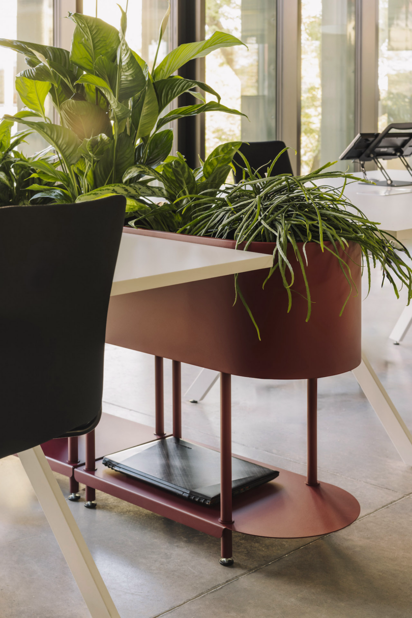 Office space with a white desk, red planter with green plants, a black chair, and a closed laptop on a shelf. Large windows in the background