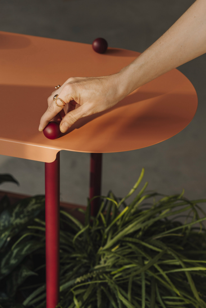 A hand touches a small red sphere on a round orange table with three red legs, positioned above green plants