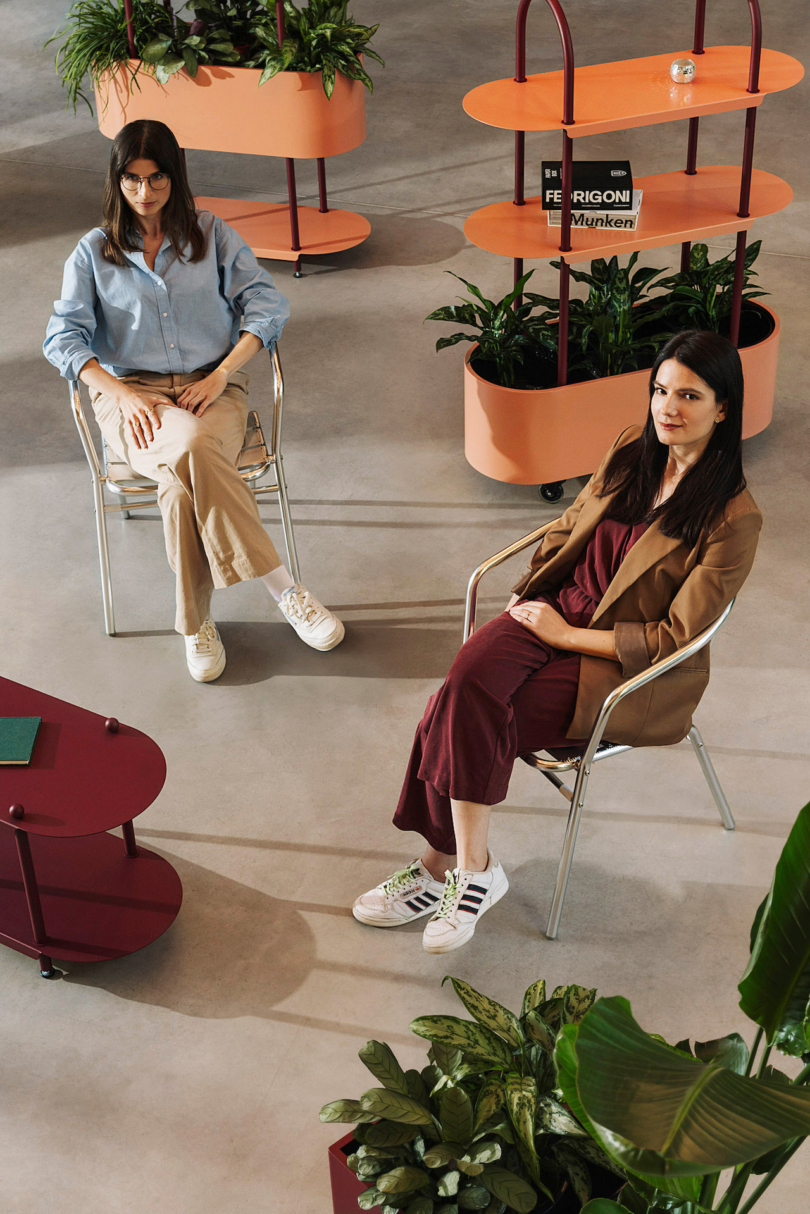 Two women sit on chairs in a modern, plant-filled space. One wears a blue shirt and beige pants; the other wears a brown blazer and maroon dress