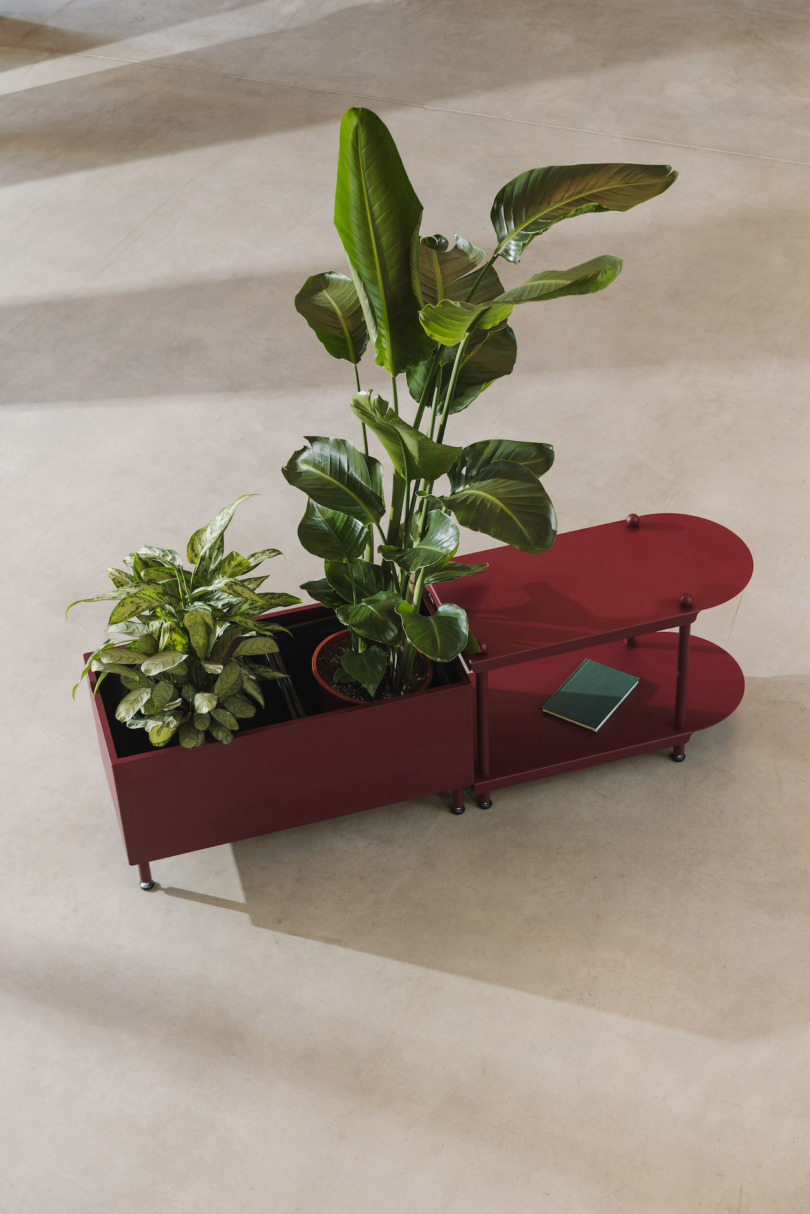 A red planter box with two green plants and a small green book on the side table, placed on a light-colored floor