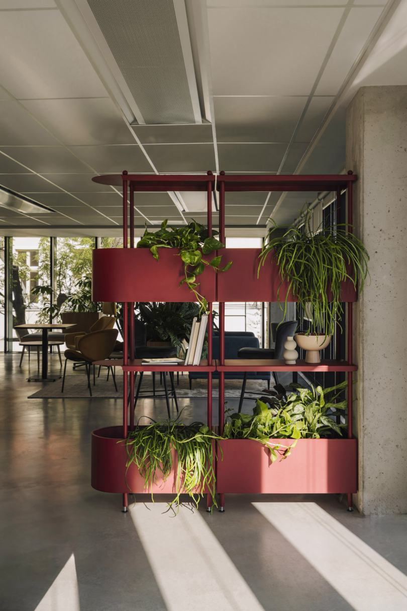 A modern indoor space featuring a red metal shelving unit with various potted green plants. Tables and chairs are visible in the background