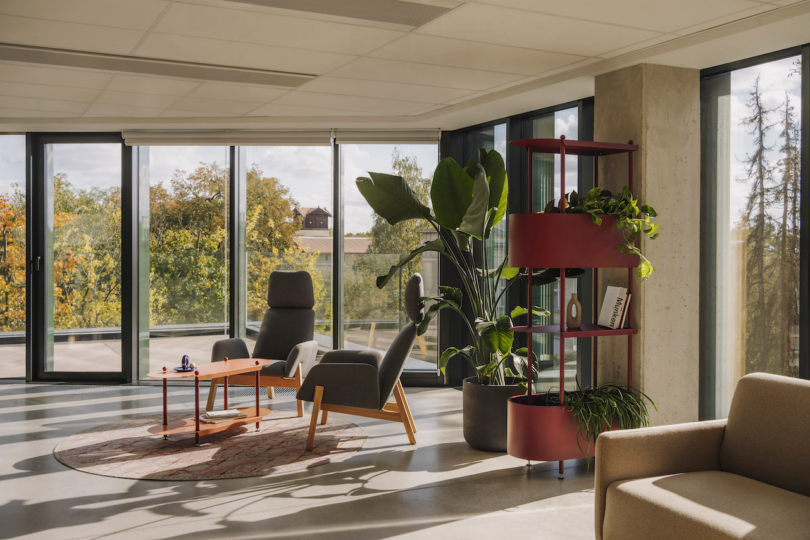 A modern, sunlit office lounge with large windows, featuring gray chairs, a small wooden table, green plants, and a red shelving unit