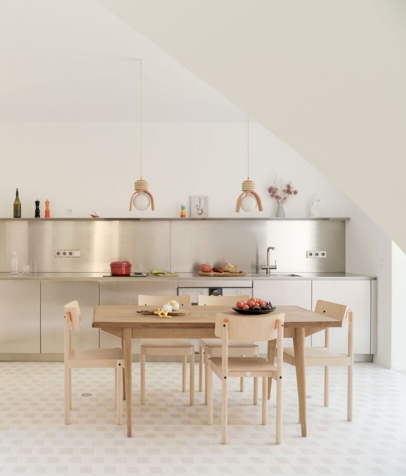 Minimalist kitchen with a wooden table and four chairs. Stainless steel cabinets and countertops, hanging pendant lights, and various kitchen items are visible.