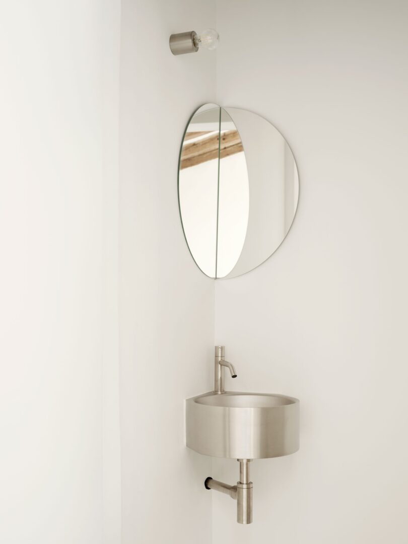 Minimalist bathroom corner with a round mirror, stainless steel sink, and modern faucet. Single bulb fixture above. White walls.