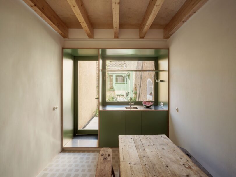 Minimalist kitchen with wooden beams, a rustic table, and green cabinets. A window and door offer natural light and garden view.