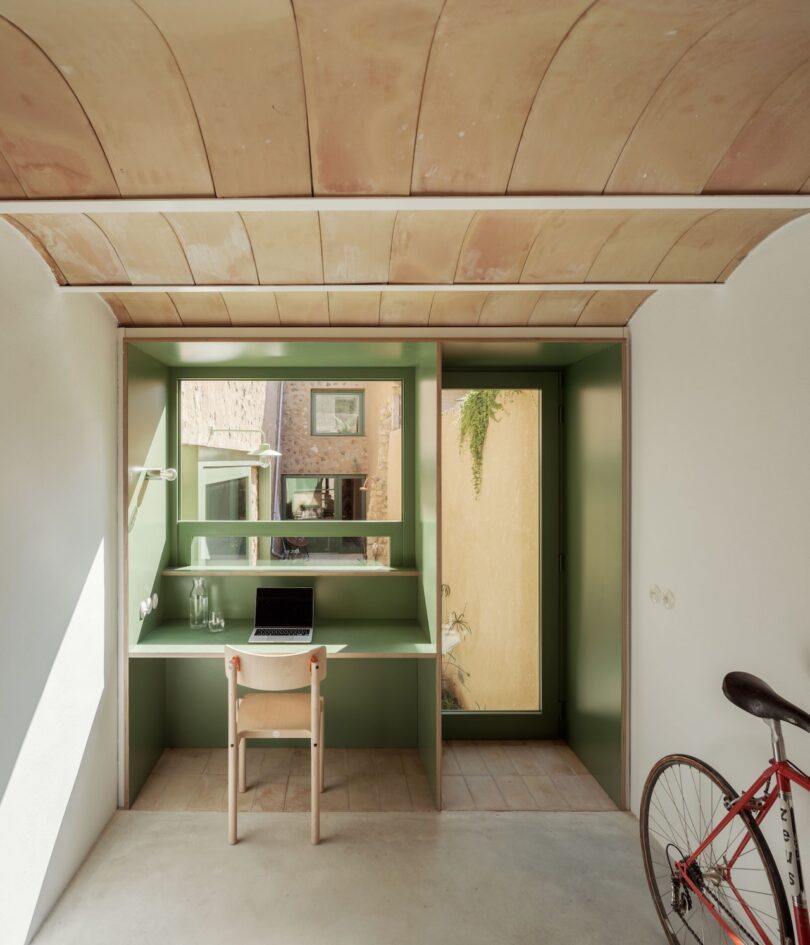 Modern workspace with a green built-in desk, chair, and a laptop. There's a series of windows, a door to the right, and a red bicycle partly visible in the foreground.