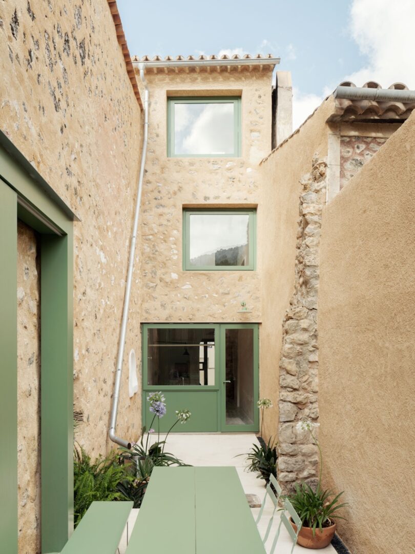 Narrow courtyard flanked by stone walls, featuring a long green table and chairs, with a two-story building in the background, green-framed windows, and plants along the sides.