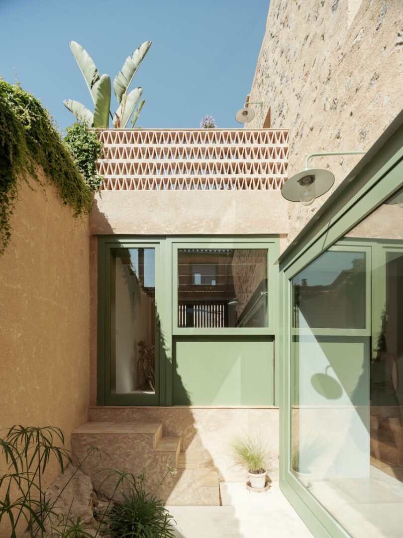 Courtyard with green-framed windows and doors, beige stone walls, lattice brick pattern, outdoor light fixture, and plants against a clear blue sky.