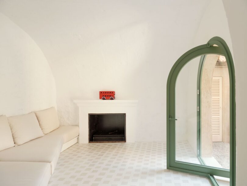 Minimalist room with white walls and arched ceiling. Beige couch along the wall, green arched door open, white fireplace with a red toy bus on the mantel, checkered floor.