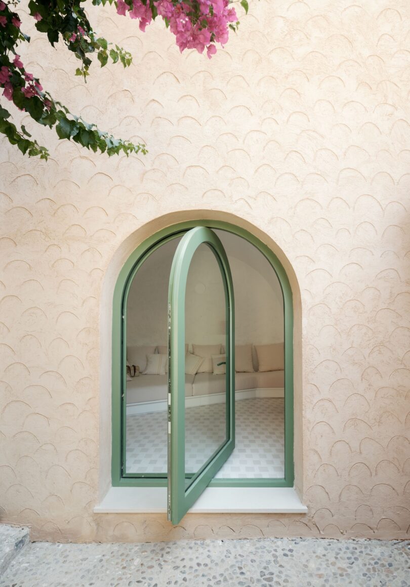 Arched window with green frame on a textured beige wall; glimpse of an interior featuring a curved sofa. Pink flowers hang in the top left corner.