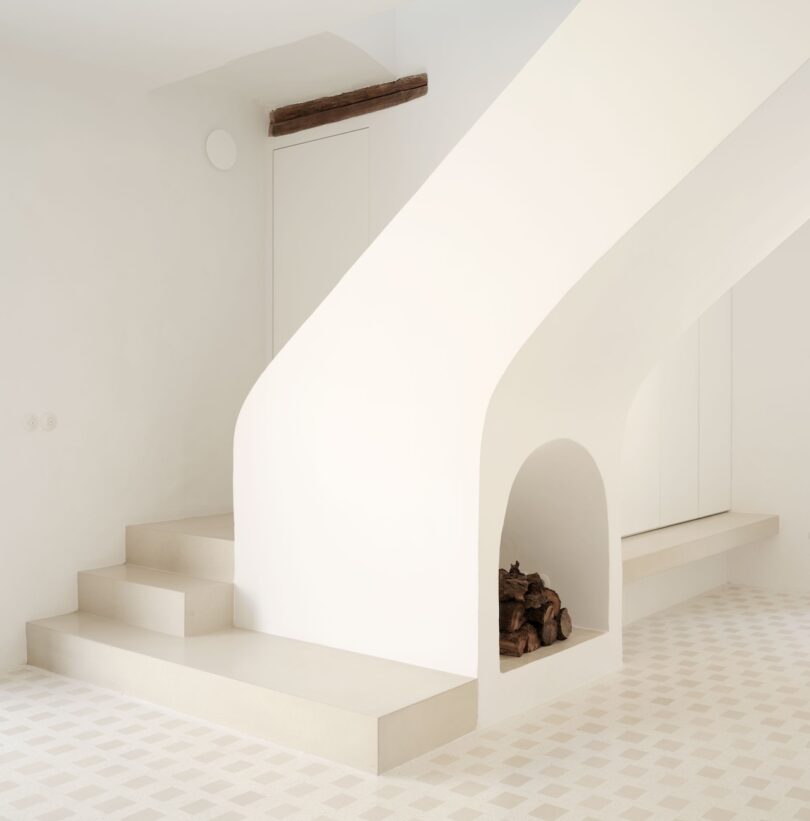 Minimalist interior with white staircase, beige steps, and a small alcove containing stacked firewood. Walls and flooring are light-colored, creating a clean and serene atmosphere.