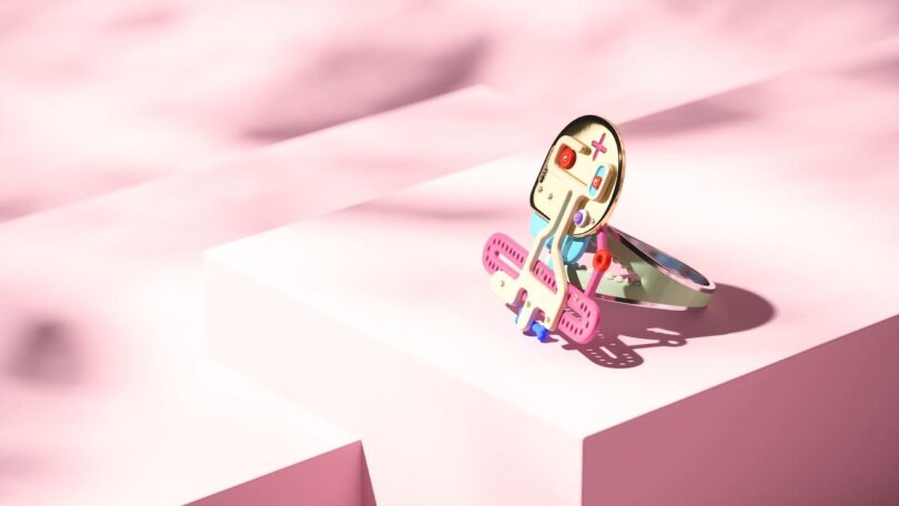 A colorful, abstract bracelet with a humanoid face sits on a pink surface against a soft pink background.