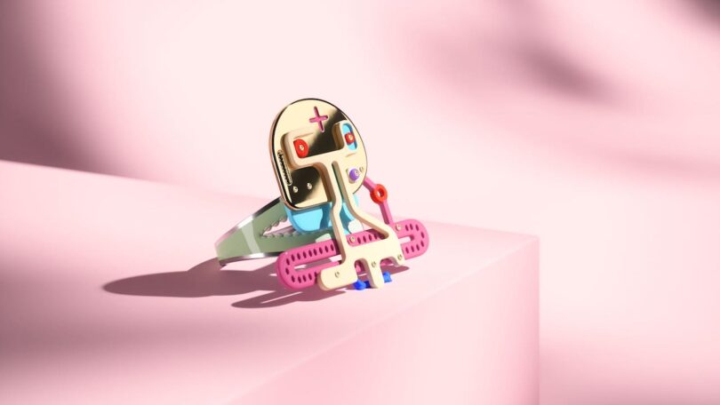 A colorful, abstract bracelet with a humanoid face sits on a pink surface against a soft pink background.