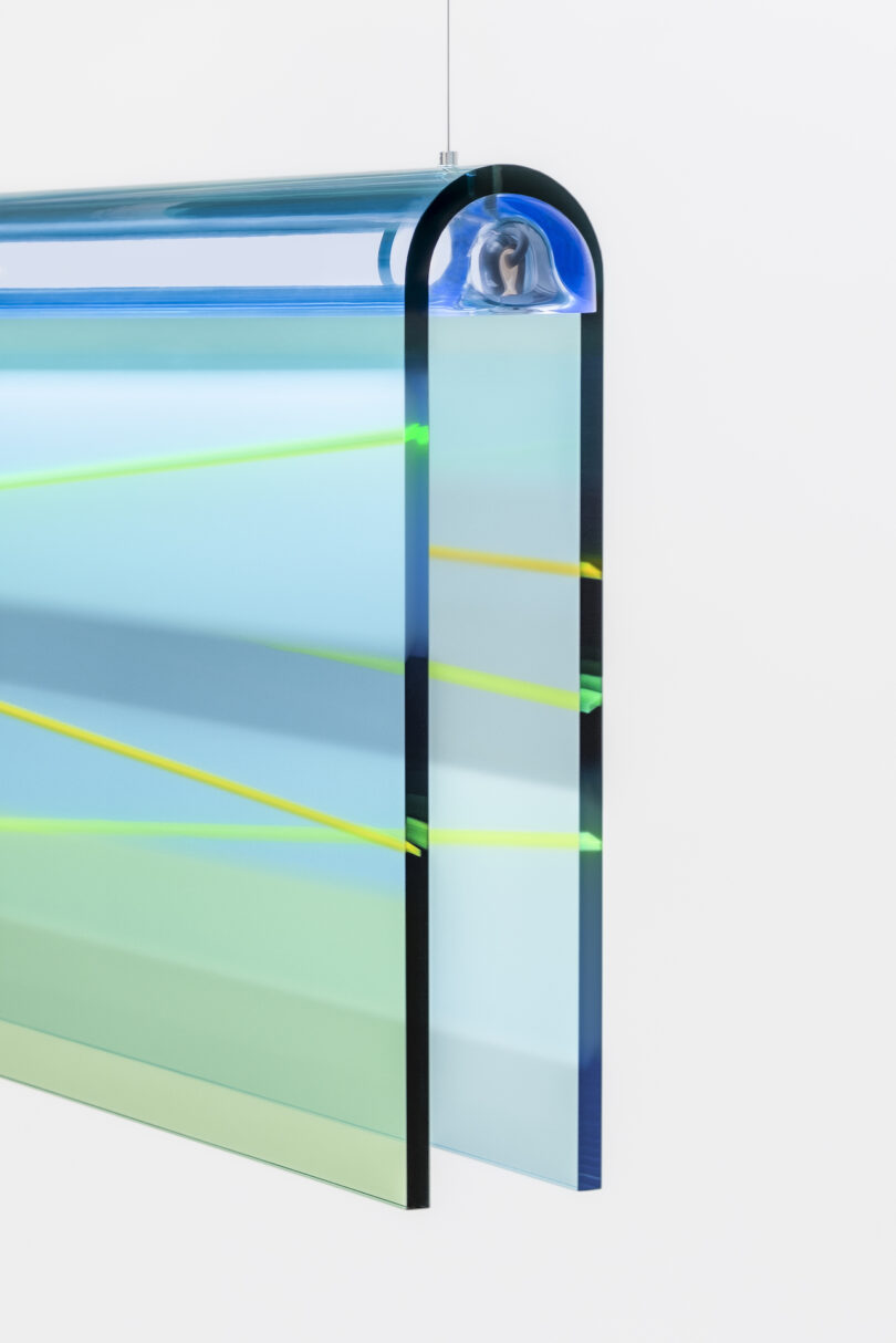 A modern art sculpture featuring transparent, layered panels with blue and green hues, adorned with diagonal yellow lines, and a curved top section