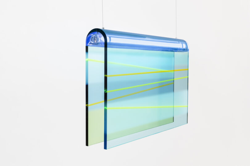 A transparent lucite pendant lamp features blue and green panels with yellow diagonal lines, suspended from the ceiling against a white background