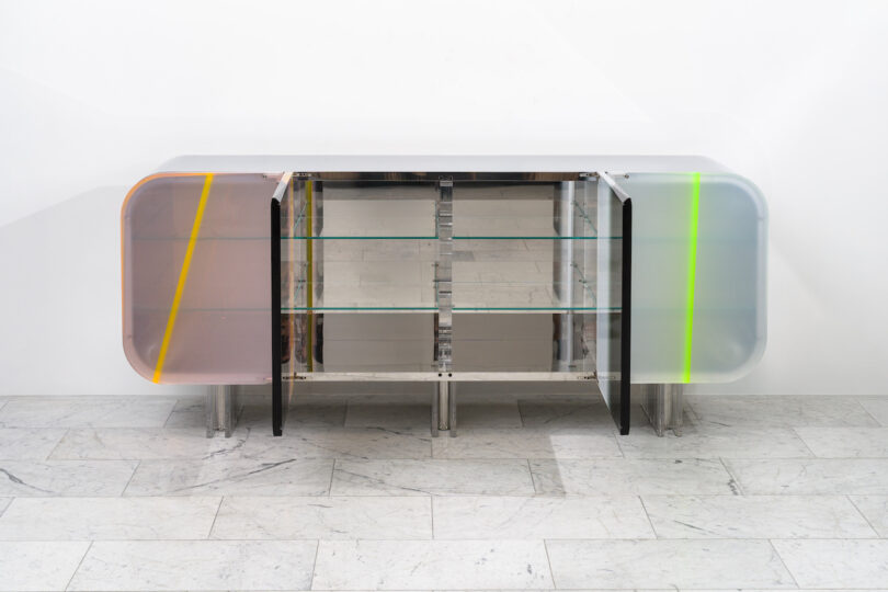 A modern sideboard with metal and lucite details, featuring partially open doors and multicolored accents, stands on a marble floor against a white wall