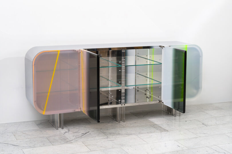 A modern, transparent cabinet with shelves and colorful doors in pink, black, and green, standing on a marble floor.