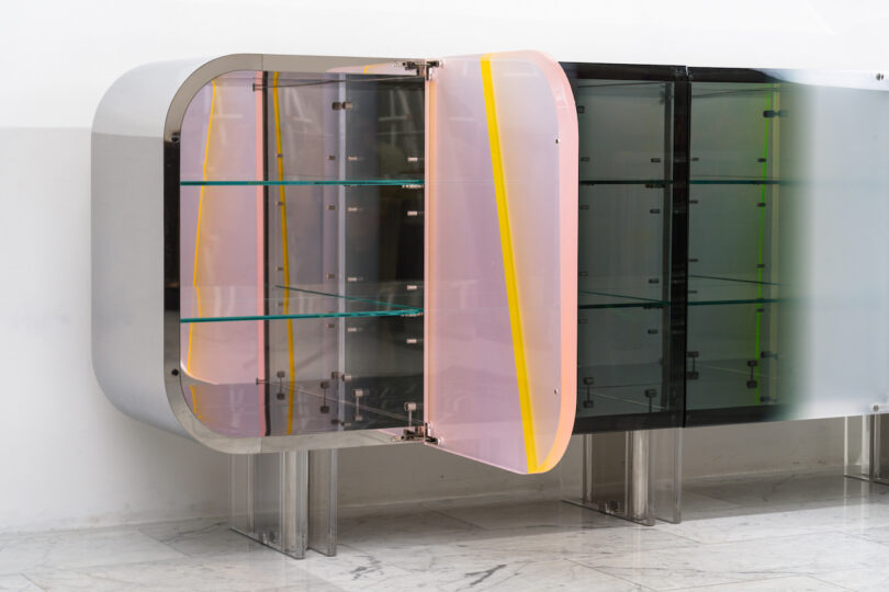 A modern lucite cabinet with rounded edges, featuring transparent pink and yellow-tinted doors. The cabinet is set against a white wall on a marble floor