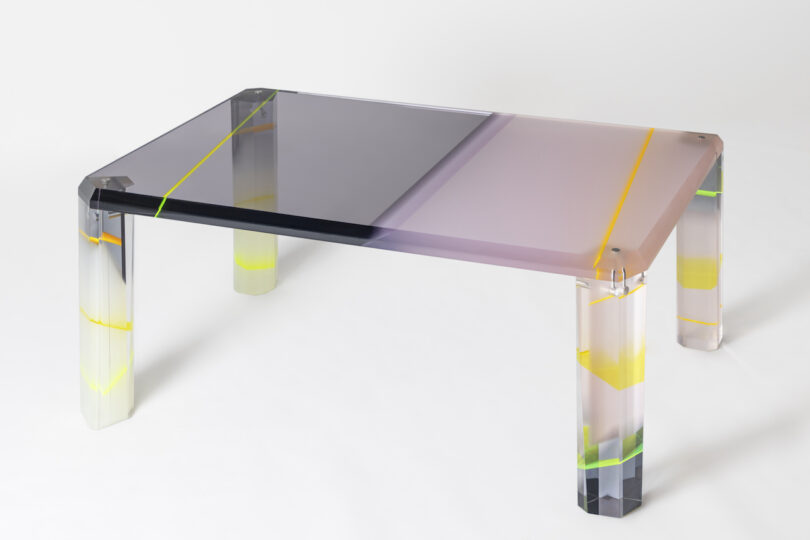 A modern lucite table with gradient-colored surfaces and legs, featuring shades of gray, purple, and yellow accents.