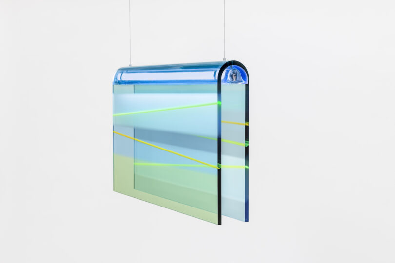 A suspended abstract lucite lamp with blue, green, and yellow hues and intersecting lines on a white background