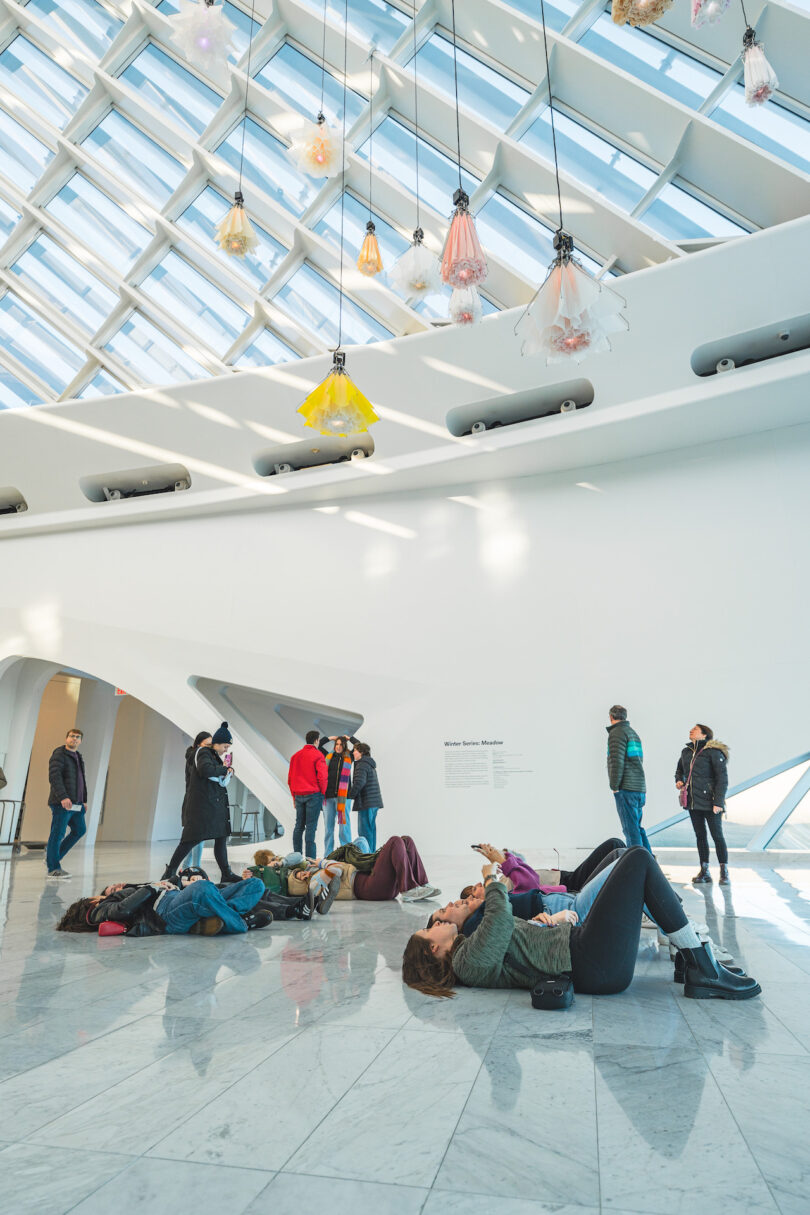 Visitors observe hanging kinetic flowers in a modern, white-walled gallery with a geometric ceiling.