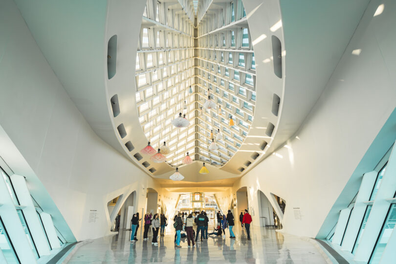 Visitors observe hanging kinetic flowers in a modern, white-walled gallery with a geometric ceiling.