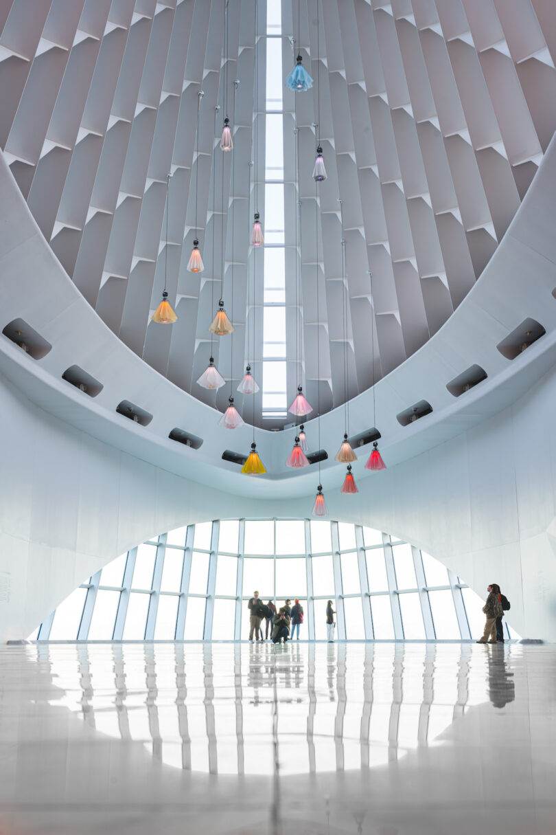 Large, modern atrium with a high ceiling and colorful, kinetic flowers. Groups of people stand near tall windows, casting reflections on the shiny floor