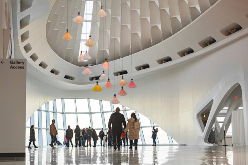 Visitors observe hanging kinetic flowers in a modern, white-walled gallery with a geometric ceiling.