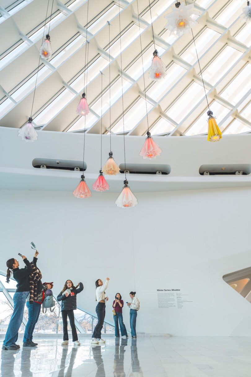 Visitors observe hanging kinetic flowers in a modern, white-walled gallery with a geometric ceiling.