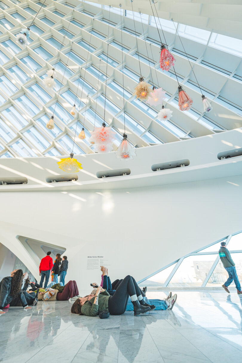 Visitors observe hanging kinetic flowers in a modern, white-walled gallery with a geometric ceiling.