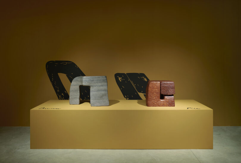 Exhibition room featuring abstract sculptural furniture on raised platforms against a yellow wall, illuminated by overhead lights