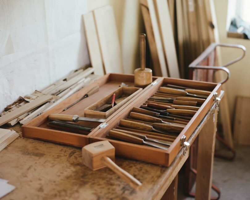 Woodworking tools neatly arranged in an open wooden box on a workbench, surrounded by planks and sketches.