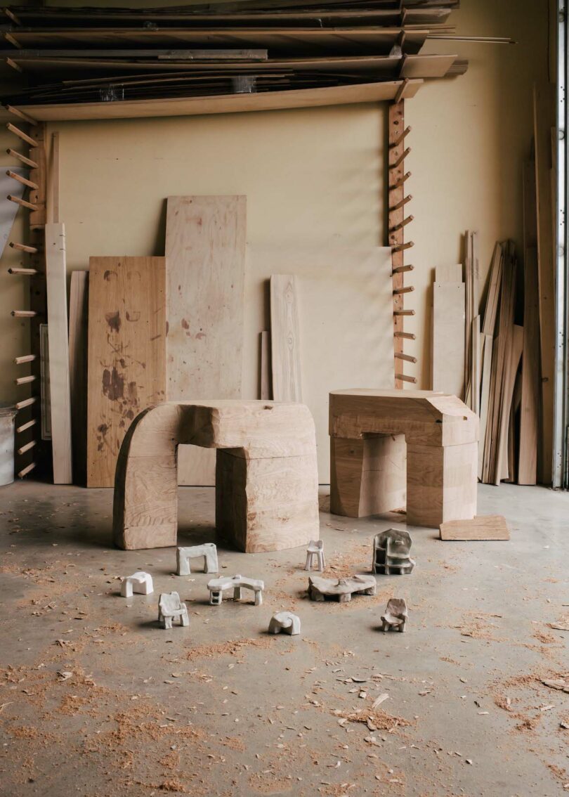 Wood workshop interior with large and small sculptures on the floor surrounded by wood shavings and stacked wooden planks against the wall