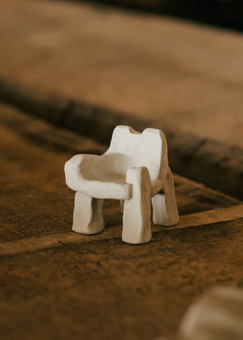 A small, abstract white clay chair with thick legs and a curved backrest is placed on a textured surface