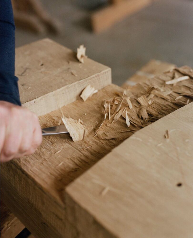 A hand uses a chisel to carve wood on a workbench, surrounded by wood shavings