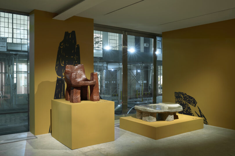 Exhibition room featuring abstract sculptural furniture on raised platforms against a yellow wall, illuminated by overhead lights