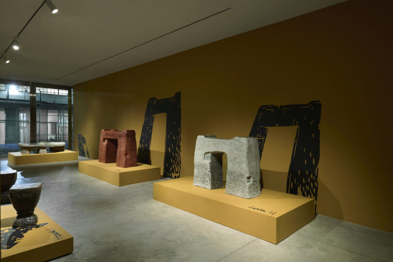 Exhibition room featuring abstract sculptural furniture on raised platforms against a yellow wall, illuminated by overhead lights