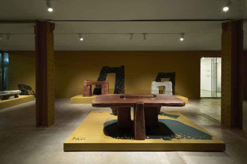 Exhibition room featuring abstract sculptural furniture on raised platforms against a yellow wall, illuminated by overhead lights