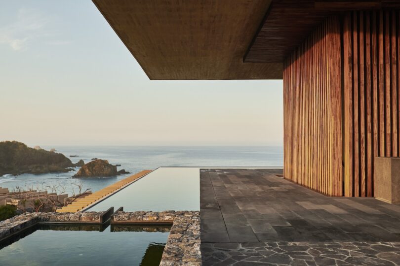 Modern building with wooden accents and stone patio overlooking an infinity pool and ocean. Rocky landscape visible in the background.