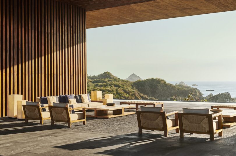 Outdoor seating area with wooden furniture on a terrace. Overlooks a scenic view of lush hills and the ocean with scattered rocks. Sunlight casts shadows on the textured wall.