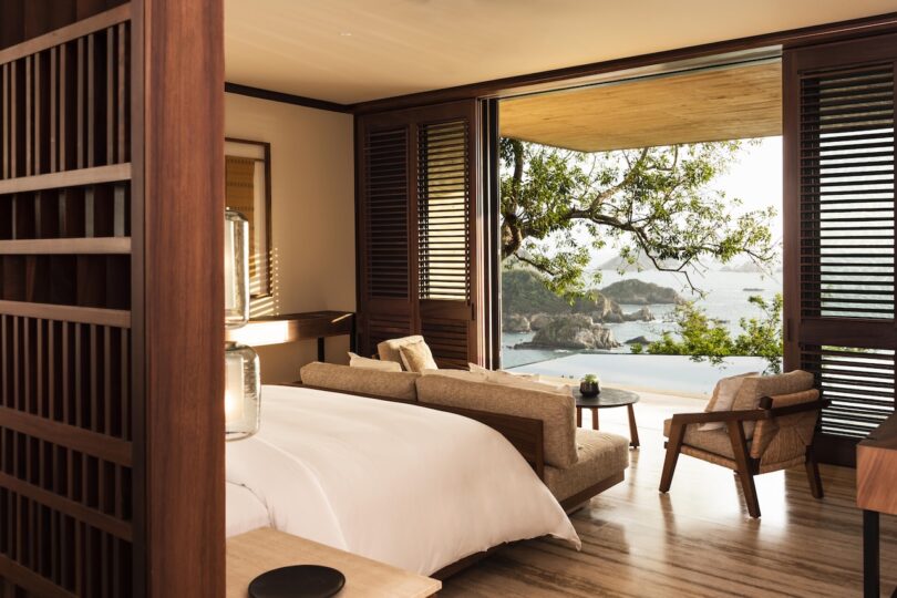 A luxurious hotel room with a large bed, wooden accents, and a view of the ocean and trees through a wide open window.