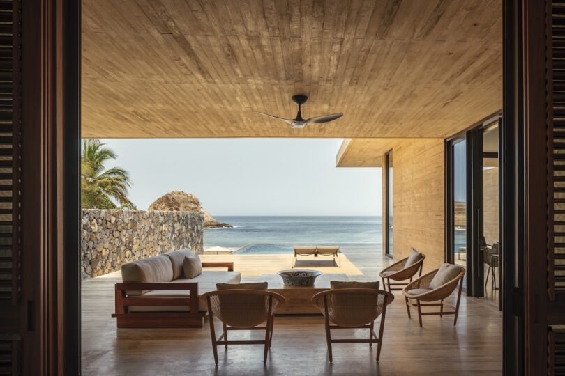 Modern open-air living space with wooden ceiling, chairs, and a sofa overlooking an ocean view. A ceiling fan and a stone wall are visible.