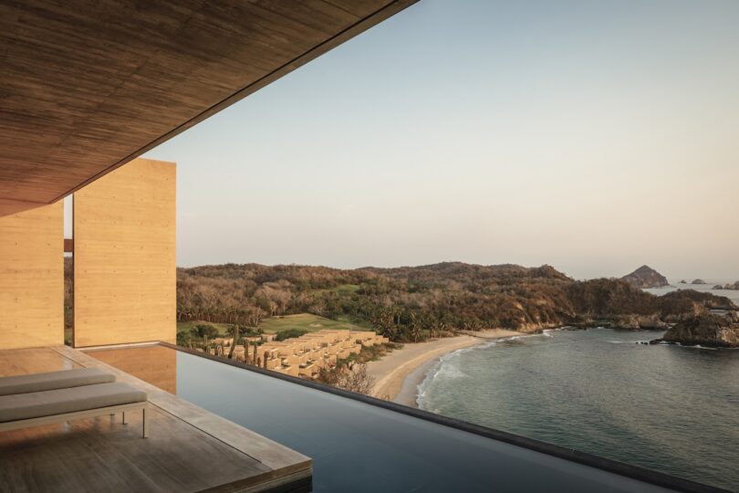 Infinity pool overlooking a sandy beach with surrounding hills and trees under a clear sky.